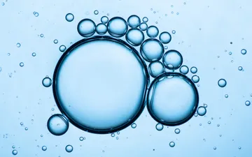 Como Aceite y Agua - Fotografía macro abstracta de gotas de aceite flotando sobre agua, formando círculos perfectos interconectados con un fondo celeste luminoso.