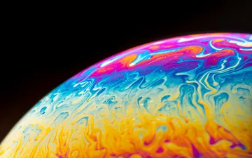 Otros Mundos - Fotografía macro abstracta de la superficie de una burbuja de jabón, mostrando patrones psicodélicos y arremolinados de colores azul, amarillo, morado y cian sobre fondo negro, asemejando un planeta gaseoso.