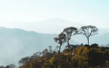 San Francisco - Paisaje brumoso del Valle Grande en Jujuy, con siluetas de montañas en diferentes planos y árboles enmarcando una pequeña casa en primer plano.